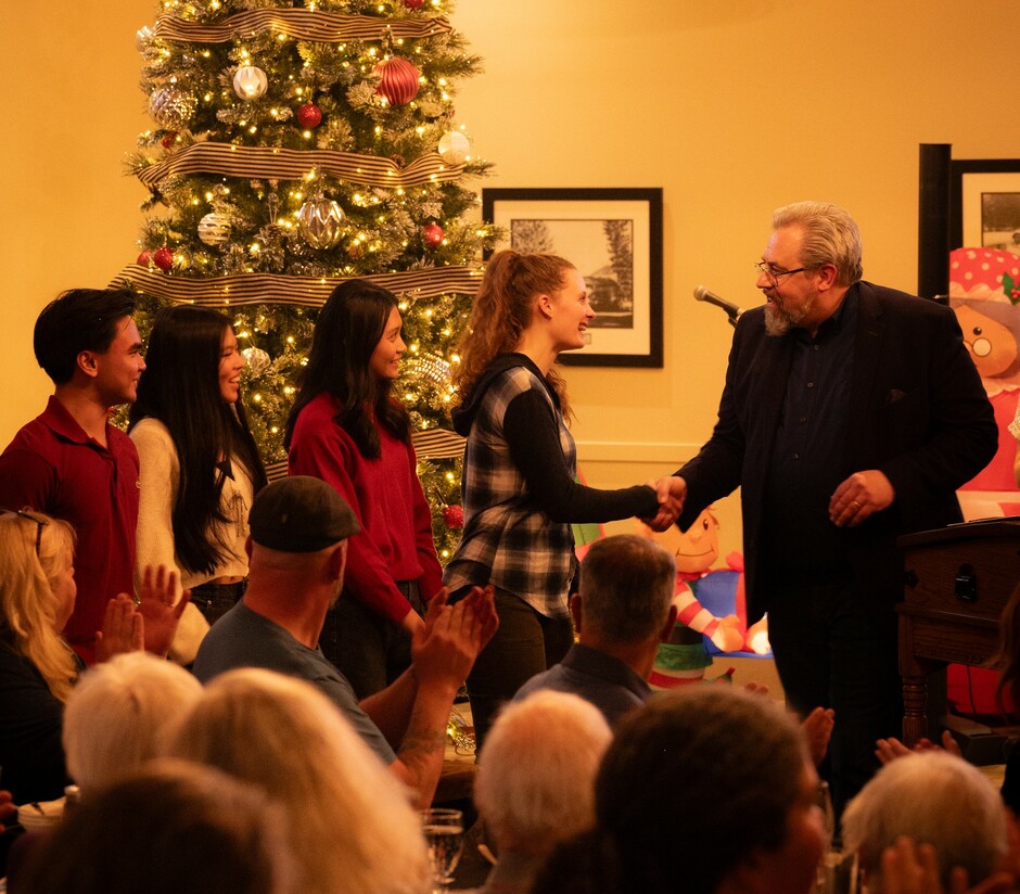 Four young volunteers stand in front of a Christmas tree, one shaking hands with Canmore's Mayor Sean Krausert, in front of a crowd